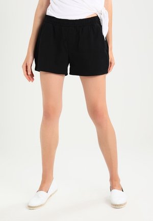 Vrouw draagt zwarte shorts met elastische taille, wit geknoopt top en witte instapschoenen, staand tegen een effen lichte achtergrond.