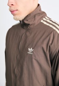 Brun zip-up jakke med høy krage, med hvite tre-stripes detaljer på skuldrene og en brodert Adidas-logo på brystet.