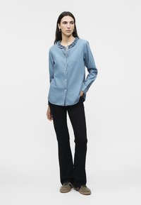 Γυναίκα που στέκεται φορώντας μπλε μπλούζα με βολάν στον γιακά, μαύρο παντελόνι και πράσινα αθλητικά παπούτσια, με τα χέρια στις τσέπες, κοιτάζοντας προς τα πλάγια.