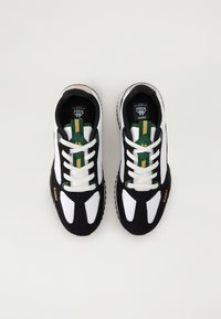 Las zapatillas en blanco y negro presentan una mezcla de materiales, con acentos verdes y amarillos en la lengüeta y los cordones, y una suela negra texturizada.