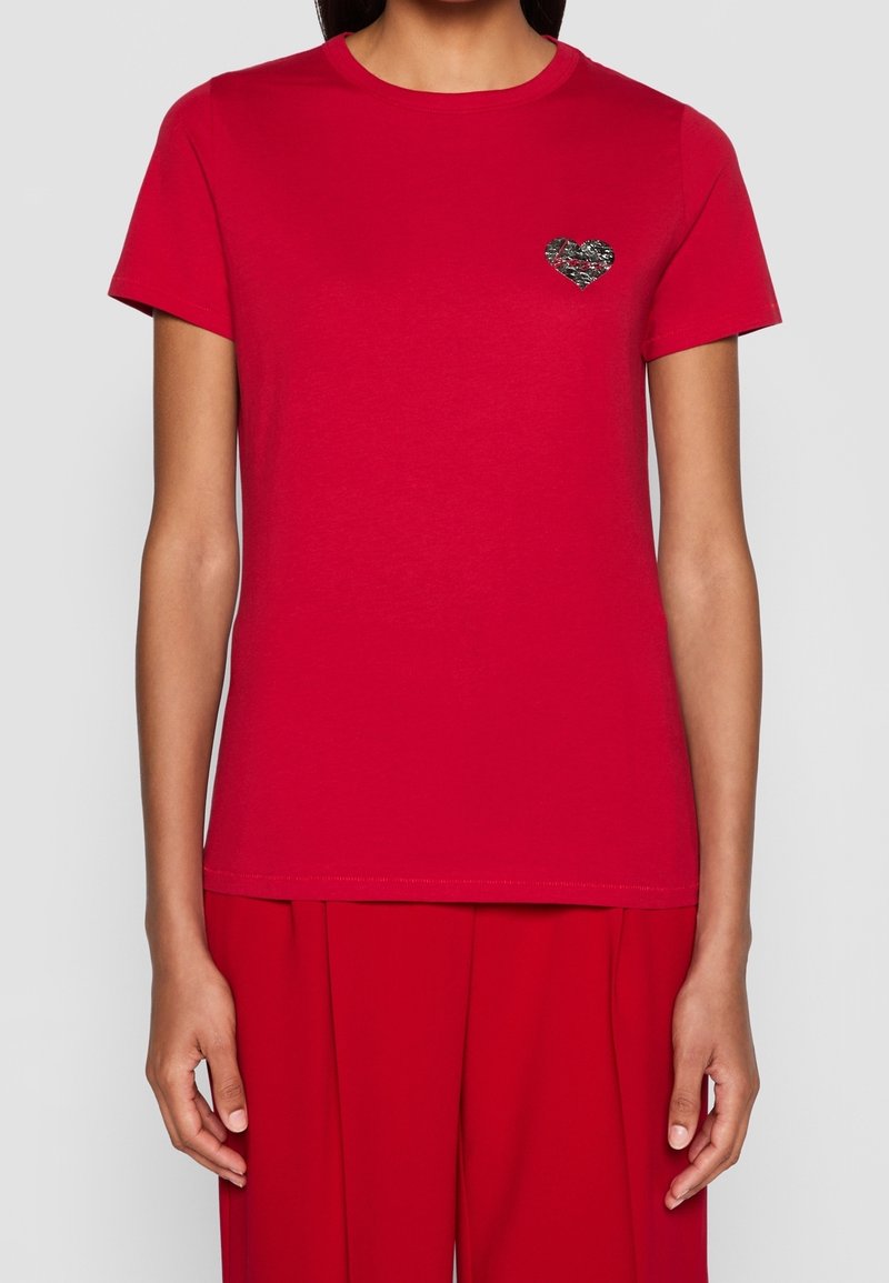 Femme portant un t-shirt rouge uni à manches courtes avec un petit motif cœur noir et blanc sur le côté gauche de la poitrine.