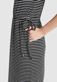 Handen vilar i fickan på en svart och vit horisontellt randig klänning med midjeband och rosett mot en neutral bakgrund.