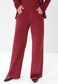 Pantaloni in corda bordeaux a gamba larga con tasche anteriori, chiusura con bottone frontale e una consistenza morbida. Abbinati a scarpe marroni a punta.