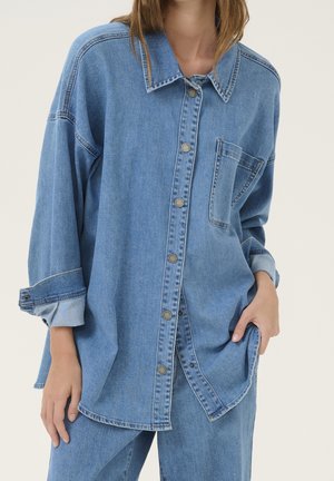 Kobieta nosząca oversize niebieską koszulę jeansową z podwiniętymi rękawami i dopasowane niebieskie dżinsy, stojąca z jedną ręką w kieszeni.
