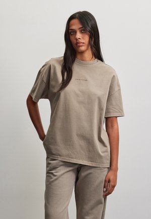 Kvinde med langt mørkt hår iført en løs beige T-shirt og matchende bukser, stående mod en ensfarvet lys baggrund.