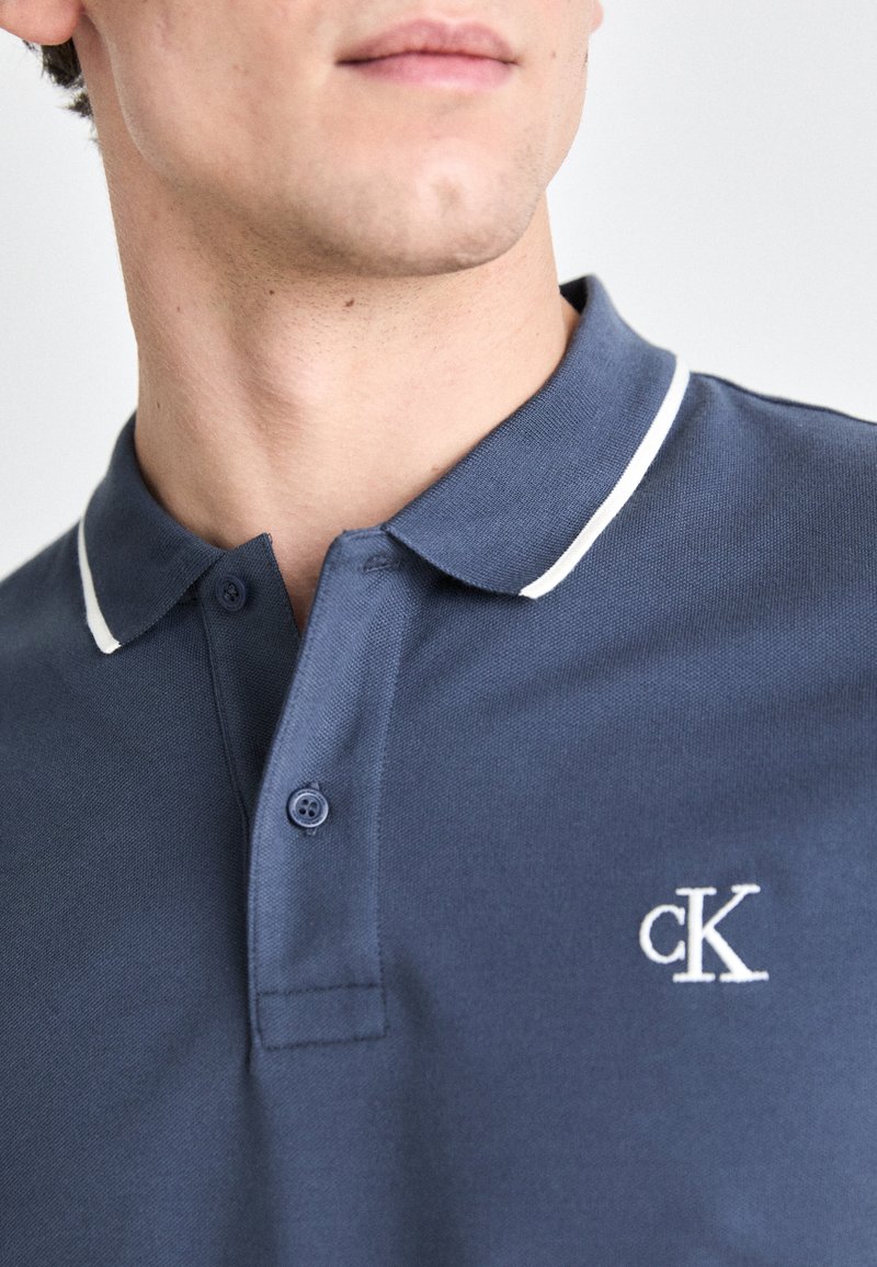 Marineblaues Poloshirt mit weißem Kragenbesatz, zwei-Knopf-Leiste, strukturiertem Stoff und kleinem gesticktem "cK"-Logo auf der Brust.