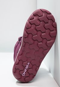 Suela de un zapato vikingo burdeos con patrón de banda de rodadura hexagonal, en ángulo contra paredes y superficie blancas.