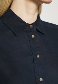 Camicia button-up blu scuro con colletto a punta, caratterizzata da bottoni in ottone e un tessuto testurizzato. Vista ravvicinata che mostra i dettagli delle cuciture.