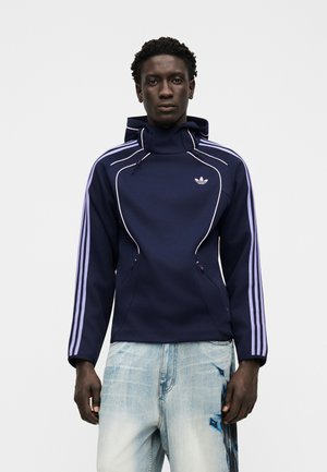 Homme portant un sweat à capuche Adidas bleu marine avec des rayures violet clair sur les manches et un jean bleu clair avec des éclaboussures de peinture bleu foncé sur la cuisse droite.