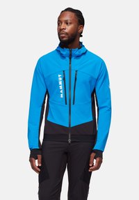 Niebieska kurtka softshell z czarnymi akcentami, z zamkiem z przodu, regulowanym kapturem i dwoma kieszeniami na piersi. Elastyczny, lekki materiał.