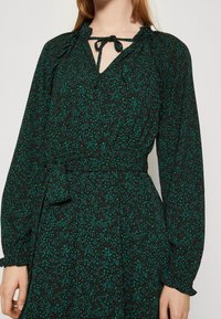 Svart klänning med grönt prickigt mönster, V-ringning med knytband, långa ärmar och skärp i midjan. Mjuk tyg med lätt glans.