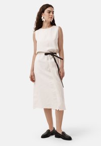 Weißes Leinenkleid mit ärmellosem Design, schwarzem Gürtel, seitlichen Schlitzen und strukturierten schwarzen Schuhen. Das Model trägt florale Ohrringe.