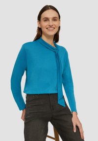 Türkise Bluse mit langen Ärmeln und Schleifen-Ausschnitt sowie glatter Textur, kombiniert mit dunklen Jeans. Modell sitzt mit Händen in den Hüften.