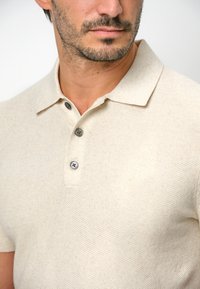 Beiges Poloshirt mit kurzen Ärmeln aus strukturiertem Stoff, mit einem flachen Kragen und drei dunklen Knöpfen an der Blende.