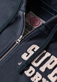 Felpa con zip di colore blu navy, caratterizzata da una zip argentata, un cordino verde e testo ricamato in rilievo. Tessuto morbido con una vestibilità rilassata.