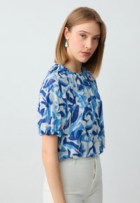 Blusa blu e bianca con scollo arricciato e maniche a puff corte, caratterizzata da un motivo a foglia in diverse tonalità di blu.