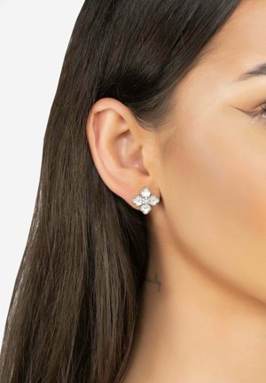 Silver floral-shaped earrings with four white stones and a central gem, set against long dark hair and a neutral background.