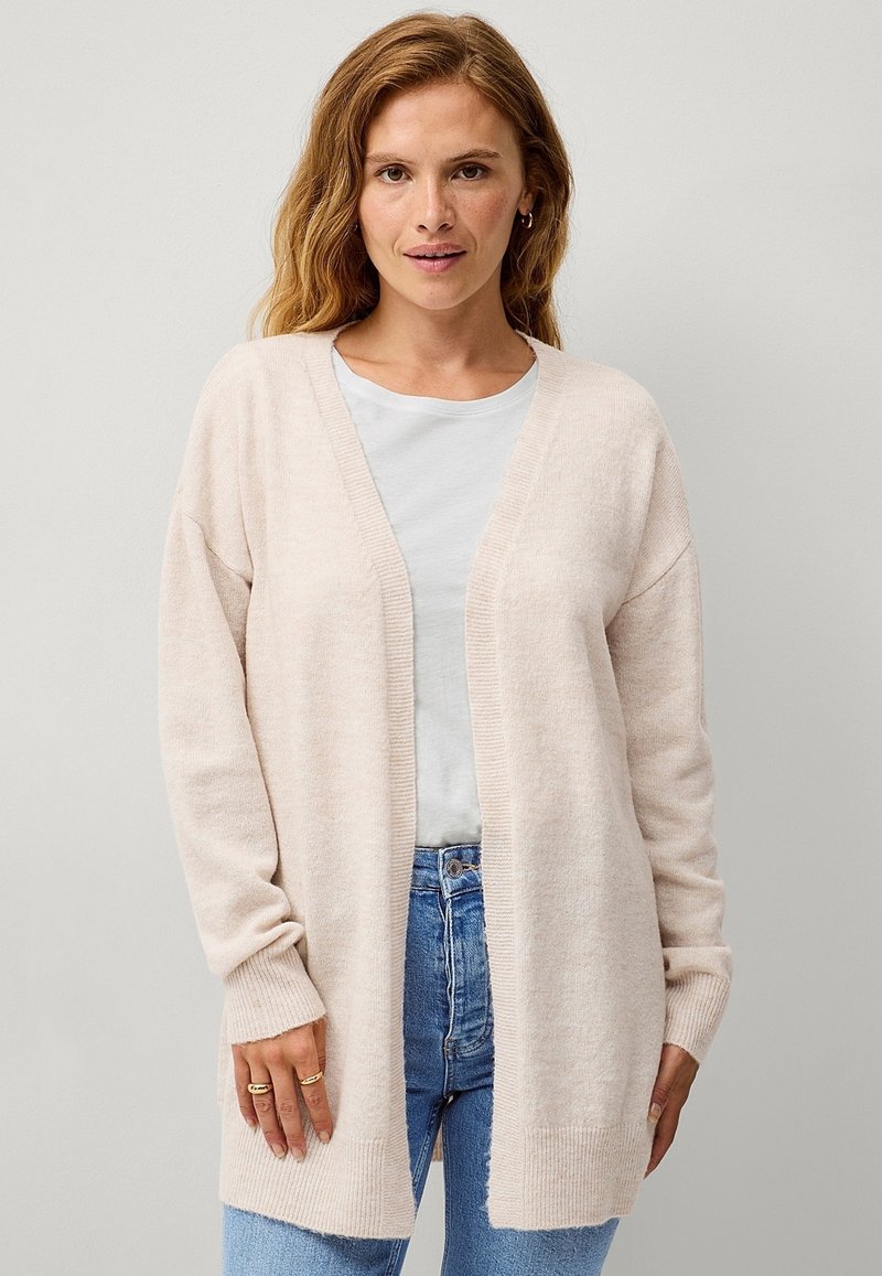 Cardigan rosa chiaro, con apertura frontale, polsini e orlo a coste, texture morbida, indossato sopra una t-shirt grigia chiaro, abbinato a jeans blu.