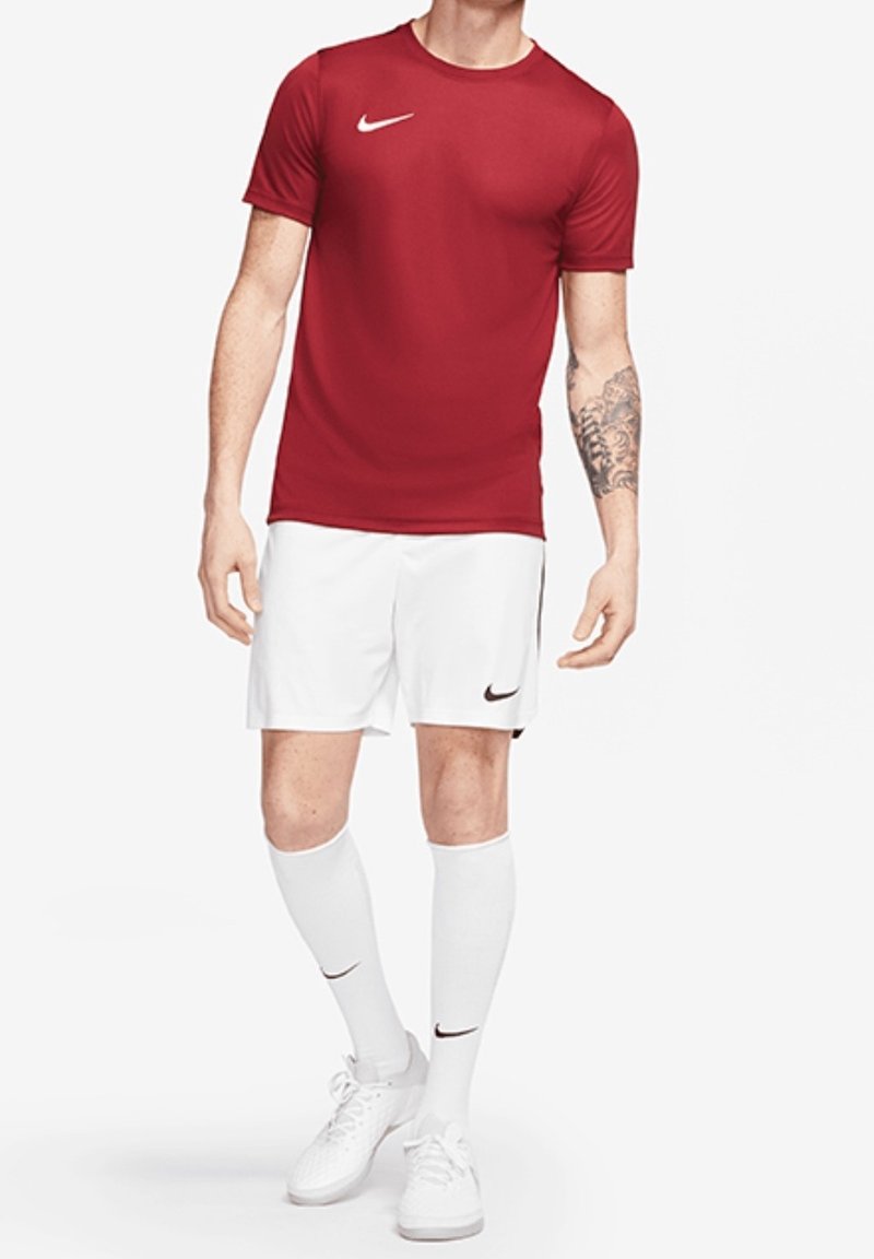 Homme portant une chemise rouge à manches courtes, un short blanc, des chaussettes blanches hautes et des baskets blanches, debout devant un fond uni.