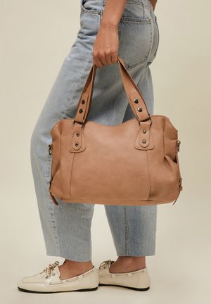 Personne portant un jean bleu clair et des mocassins crème tenant un grand sac à main en cuir marron clair par ses poignées.