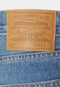 Étiquette en cuir marron avec texte et motifs embossés, cousue sur un tissu en denim bleu, présentant un design classique à rivets.