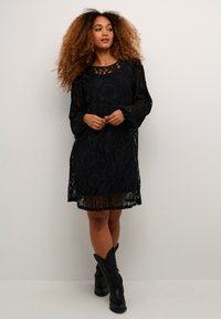 Rochie neagră din dantelă cu mâneci lungi, transparente, model floral, decolteu rotund și lungime peste genunchi. Asortată cu cizme negre de tip cowboy.