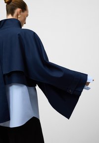 Giacca in stile mantella di colore blu navy con maniche ampie sopra una camicia azzurra chiara. Tessuto liscio, colletto a punta e polsini abbottonati visibili.