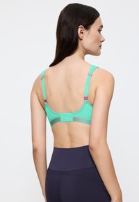 Kvinde set fra siden iført en teal sports-bh med justerbare stropper og marineblå leggings med høj talje mod en ensfarvet baggrund.