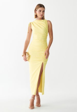 Mujer con un vestido amarillo sin mangas fruncido con abertura en el muslo, sosteniendo un bolso de mano dorado, pendientes dorados, pulsera y sandalias doradas de tiras.