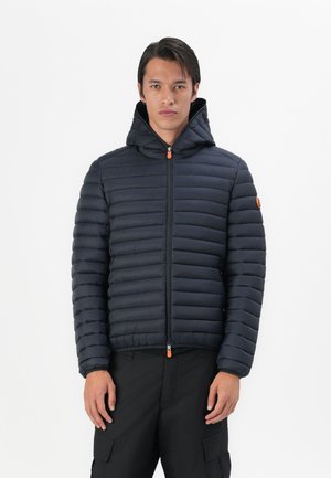Gewatteerde marineblauwe jas met capuchon en ritssluiting aan de voorkant. Voorzien van horizontale stiksels, elastische manchetten en een oranje ritslip.
