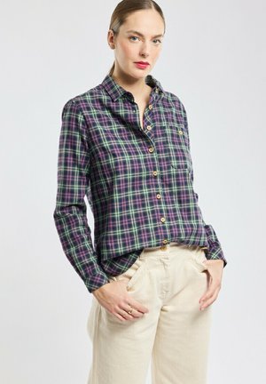 Chemise à carreaux verte et violette à boutons avec col, dotée d'une poche poitrine et de boutons en bois, associée à un pantalon beige.