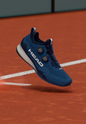 Blauwe HEAD sportschoen met BOA-sluitingen die klei opstuift op een tennisbaan vlak bij een witte lijn.