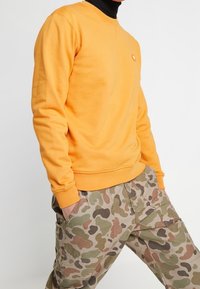 Gele sweater met ronde hals en ribgebreide manchetten, gecombineerd met camouflage cargobroek met zakken en een gestructureerde stof.