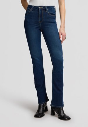 Femme portant un jean bleu foncé droit, des bottines noires à talons et un haut blanc sans manches, debout devant un fond uni.