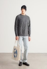 Pelēks džemperis ar adījumu, gaiši džinsi, melna cepure un melni apavi; rokās tur bēša krāsas iepirkumu somu ar zilu grafisku dizainu. Neitrāla fonā.