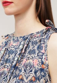 Närbild av en kvinna som bär en ärmlös blus med blommönster och knutna axelband, samt röd läppstift.