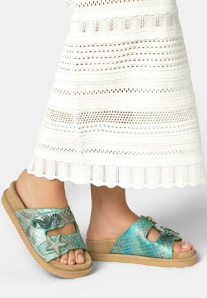 Pies con sandalias de punta abierta con un patrón de escamas turquesas y adornos con temática marina, combinados con una falda midi blanca de ojales.