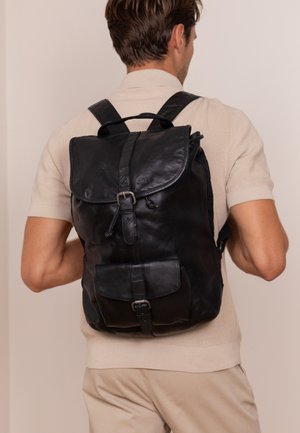 Zwarte leren rugzak met een klepafsluiting, verstelbare schouderbanden, een voorzak met een gesp en een zachte, gestructureerde afwerking.