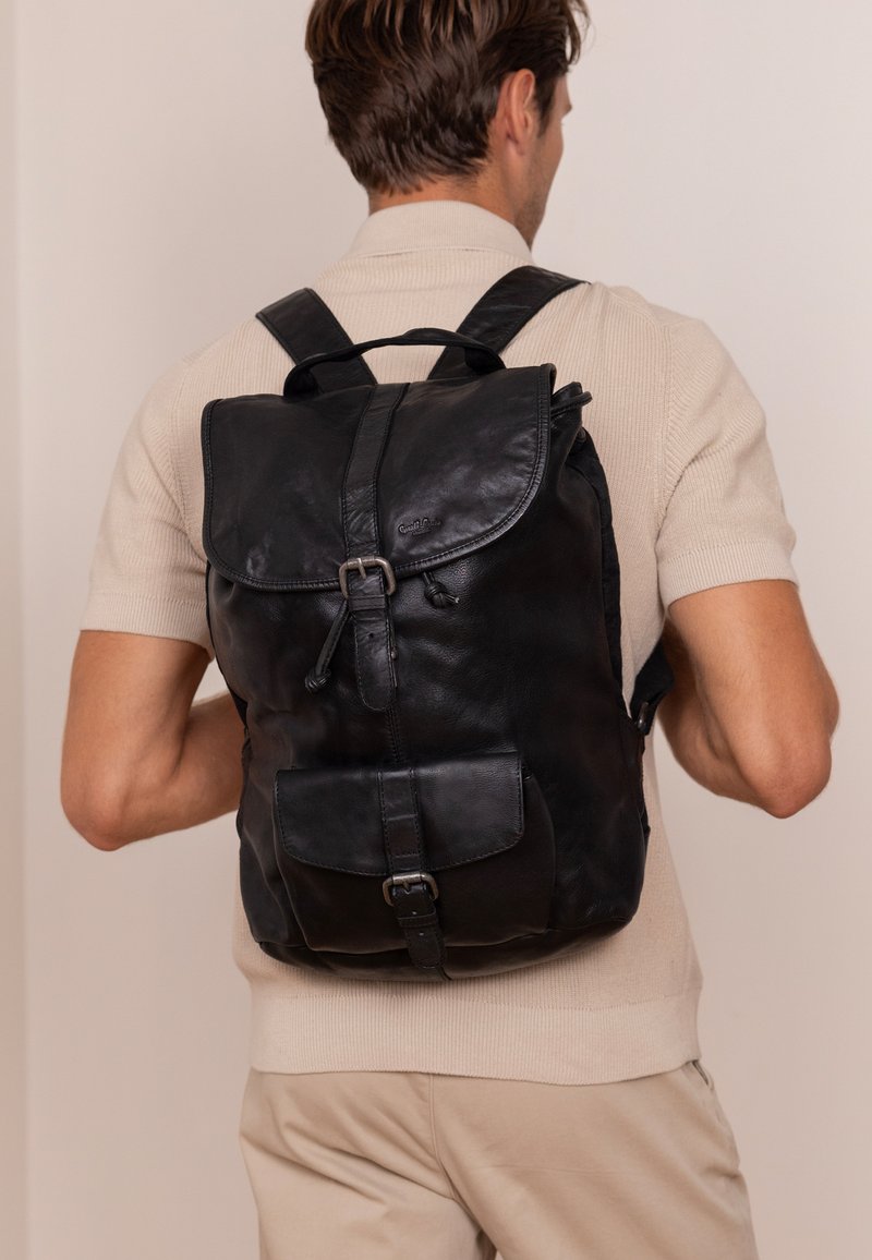 Schwarzer Leder-Rucksack mit Klappenverschluss, verstellbaren Trägern, Fronttasche mit Schnalle und weicher, texturierter Oberfläche.