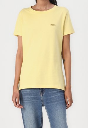 Person wearing a plain yellow short-sleeve t-shirt with "BOSS" logo on left chest and blue denim jeans, standing against white background.