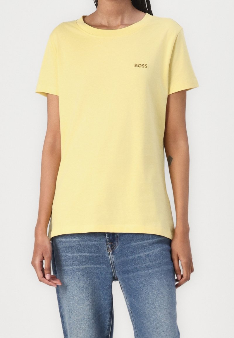 Personne portant un t-shirt jaune uni à manches courtes avec le logo « BOSS » sur la poitrine gauche et un jean en denim bleu, debout contre un fond blanc.