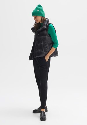 Black puffer vest with high collar, green long-sleeve shirt, black trousers, and black loafers. Green knit beanie adds accent.