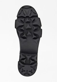 Semelle de chaussure en caoutchouc noir présentant une texture antidérapante avec des motifs angulaires et le nom de la marque "GUESS" embouti sur le côté.