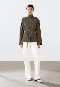 Vrouw in een olijfgroene knoopjasje met ceintuur, witte rechte broek en witte ballerina's, staand op een grijze vloer tegen een witte muur.