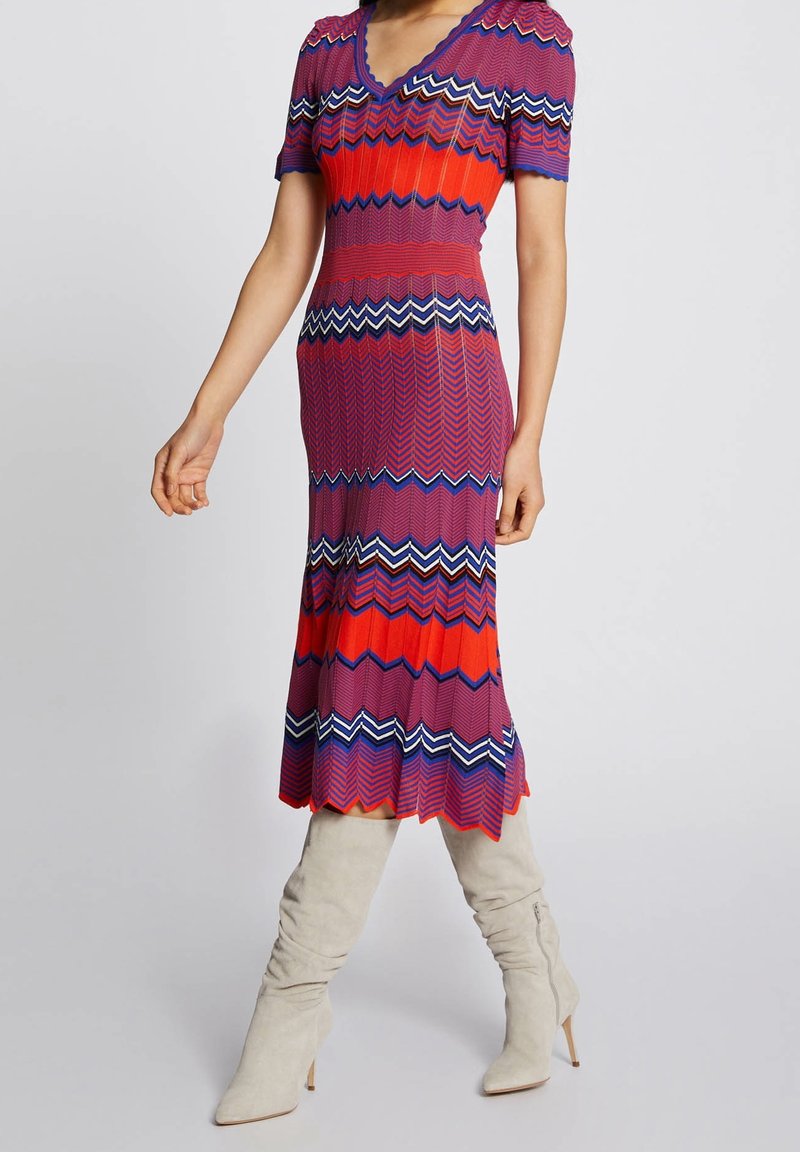Femme portant une robe ajustée à motif en zigzag, longueur genou, aux couleurs rouge, violet, blanc et bleu, avec des manches courtes et des bottes beige montantes jusqu'aux genoux.