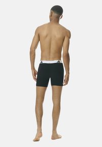 Hombre de pie descalzo mostrando la espalda, vistiendo calzoncillos negros con una cinturilla blanca que presenta los logotipos de Adidas en negro.