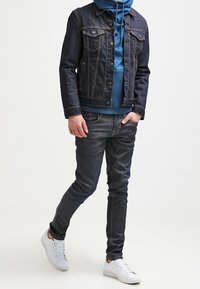 Chaqueta de mezclilla oscura con dos bolsillos en el pecho, llevada sobre un hoodie azul. Jeans oscuros ajustados y zapatillas blancas completan el conjunto.