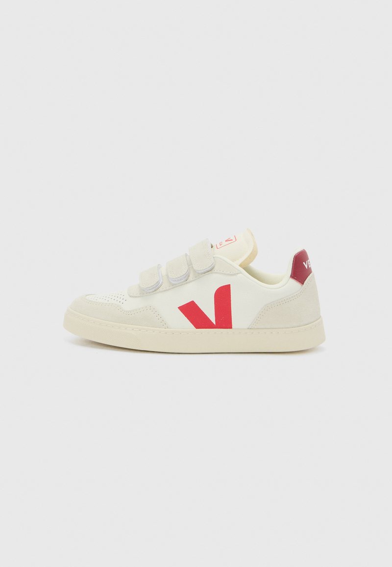 Sneaker bianca con logo rosso, cinturini in velcro, suola in gomma, dettagli in suede, perforazioni sulla punta e colletto imbottito.