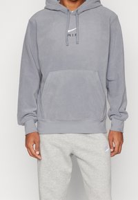 Felpa grigia in pile con tasca frontale, cappuccio regolabile e un logo a contrasto. Abbinata a pantaloni della tuta grigi chiari con un piccolo logo.