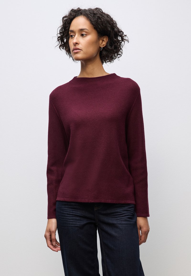 Bordeauxfarbener Strickpullover mit langen Ärmeln, Rundhalsausschnitt und strukturiertem Finish. Kombiniert mit dunkelblauen Jeans.